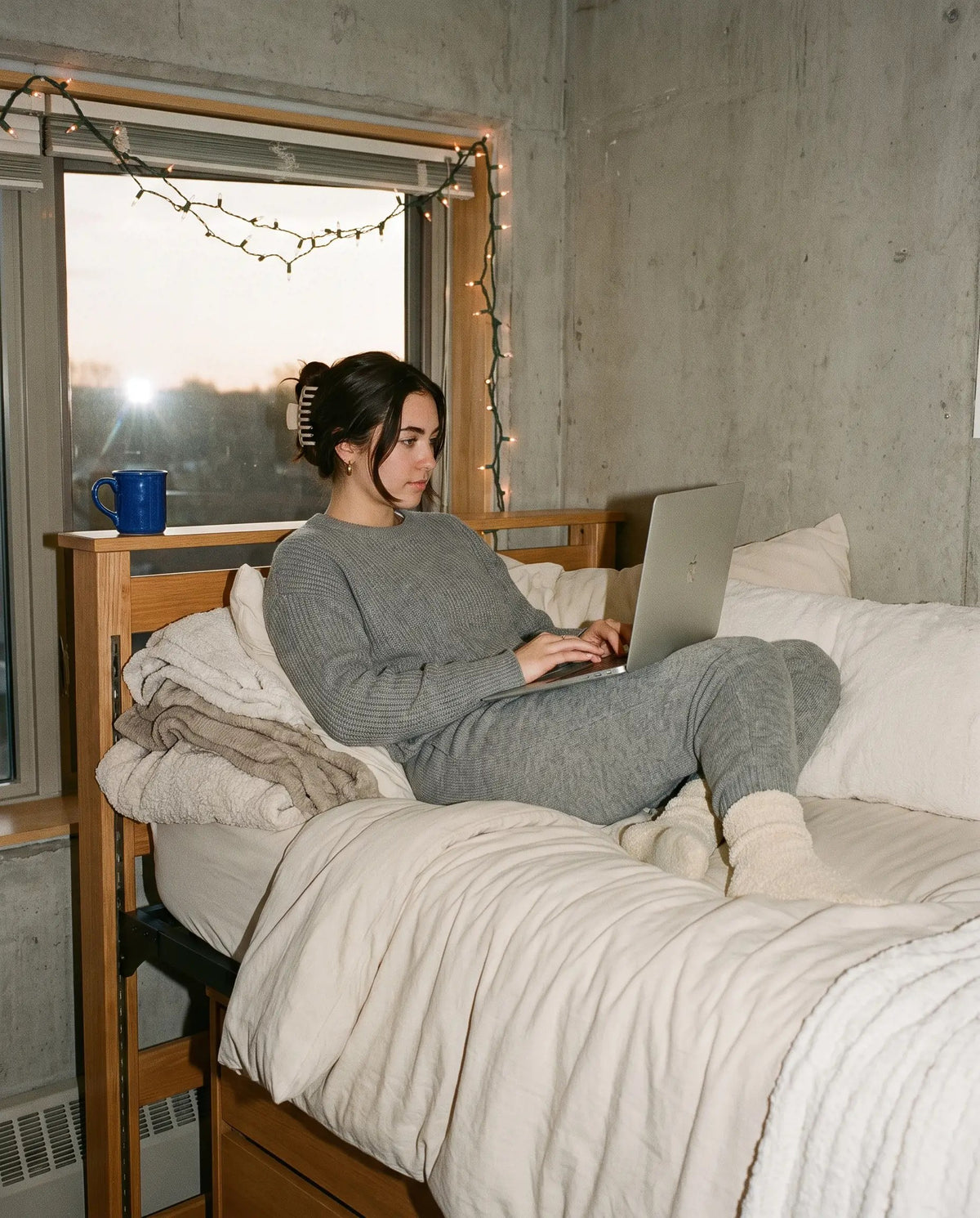 Student in a grey knit lounge set sitting on a dorm bed with a laptop, string lights on the window