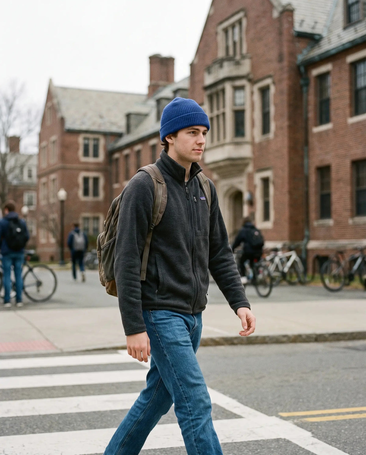 Student crossing a campus street in a black fleece, straight jeans, cobalt beanie and backpack, brick halls behind