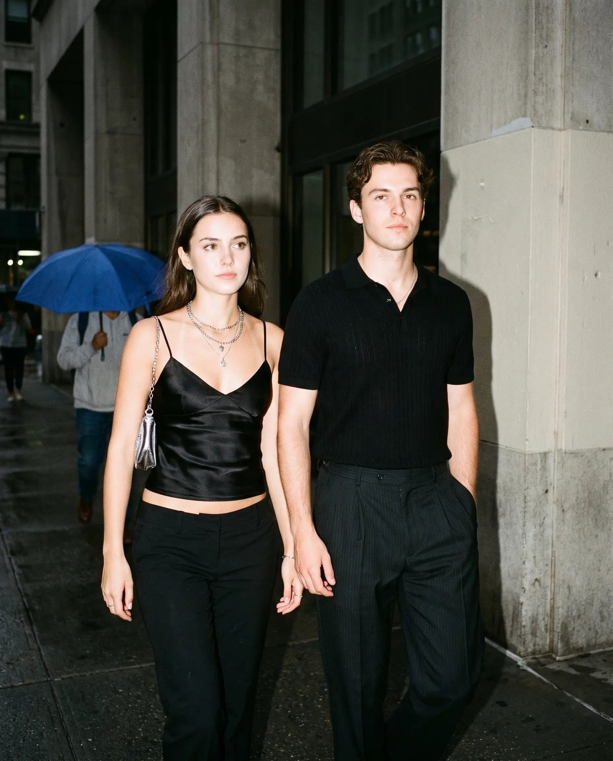 Two friends out at night by a concrete building: black satin cami with black trousers, black knit polo with pleated trousers