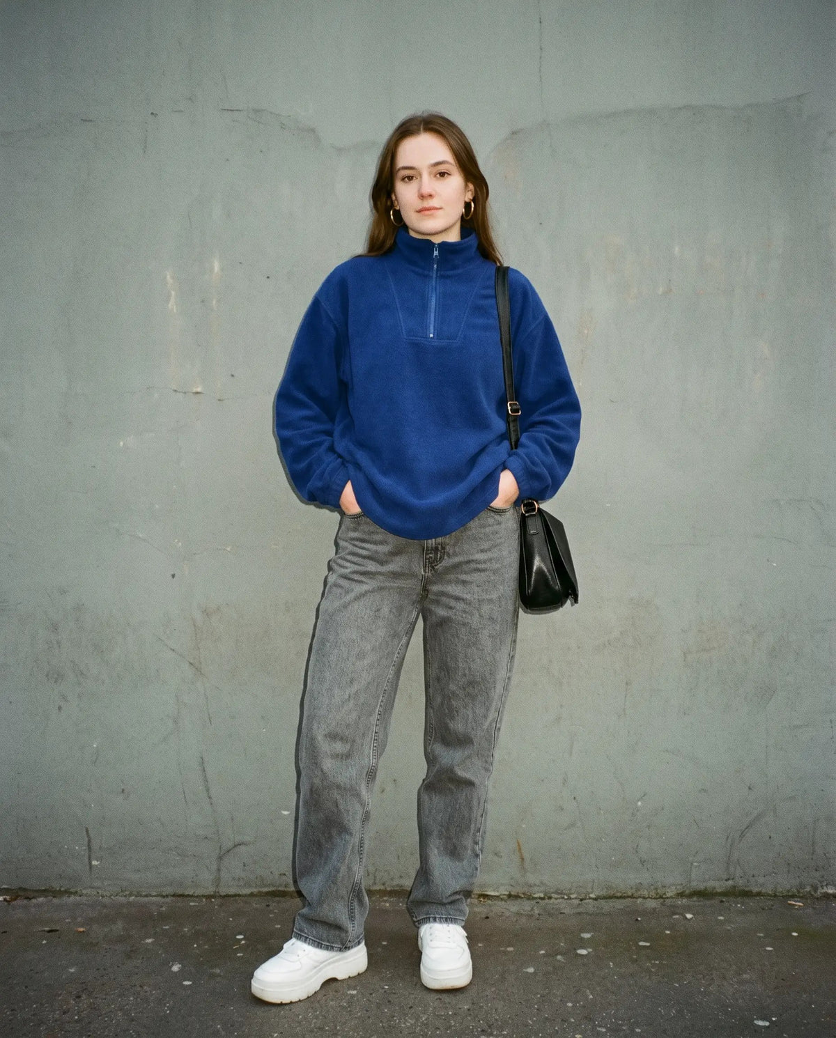 Woman in a cobalt fleece half-zip, grey wide-leg jeans, white sneakers and a small black shoulder bag against a concrete wall