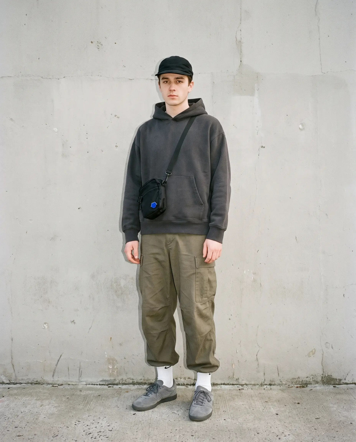 Man in a charcoal hoodie, olive cargo pants, black cap and small crossbody bag against a concrete wall