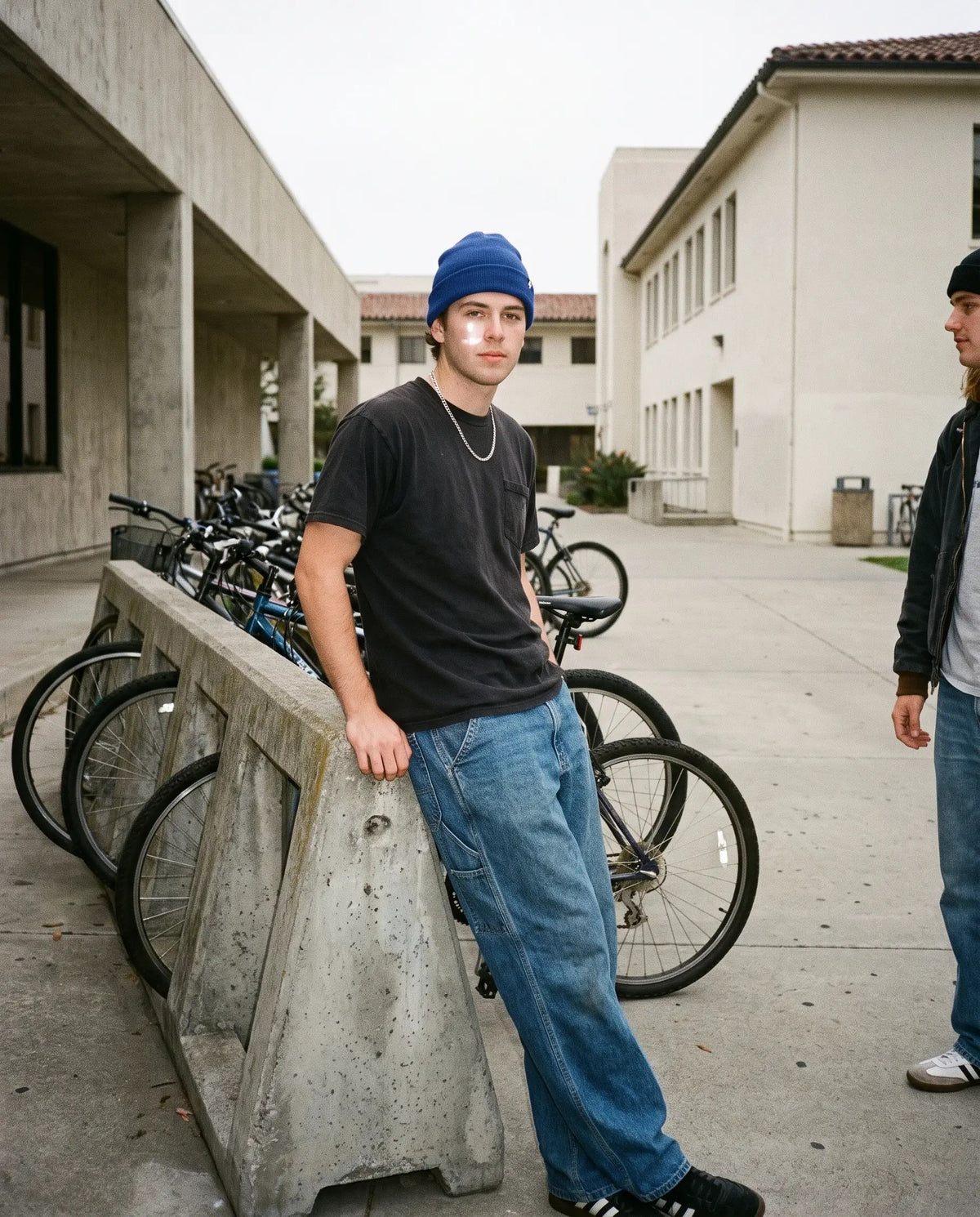 Student in a black tee, baggy light-wash jeans and cobalt beanie leaning on a campus bike rack