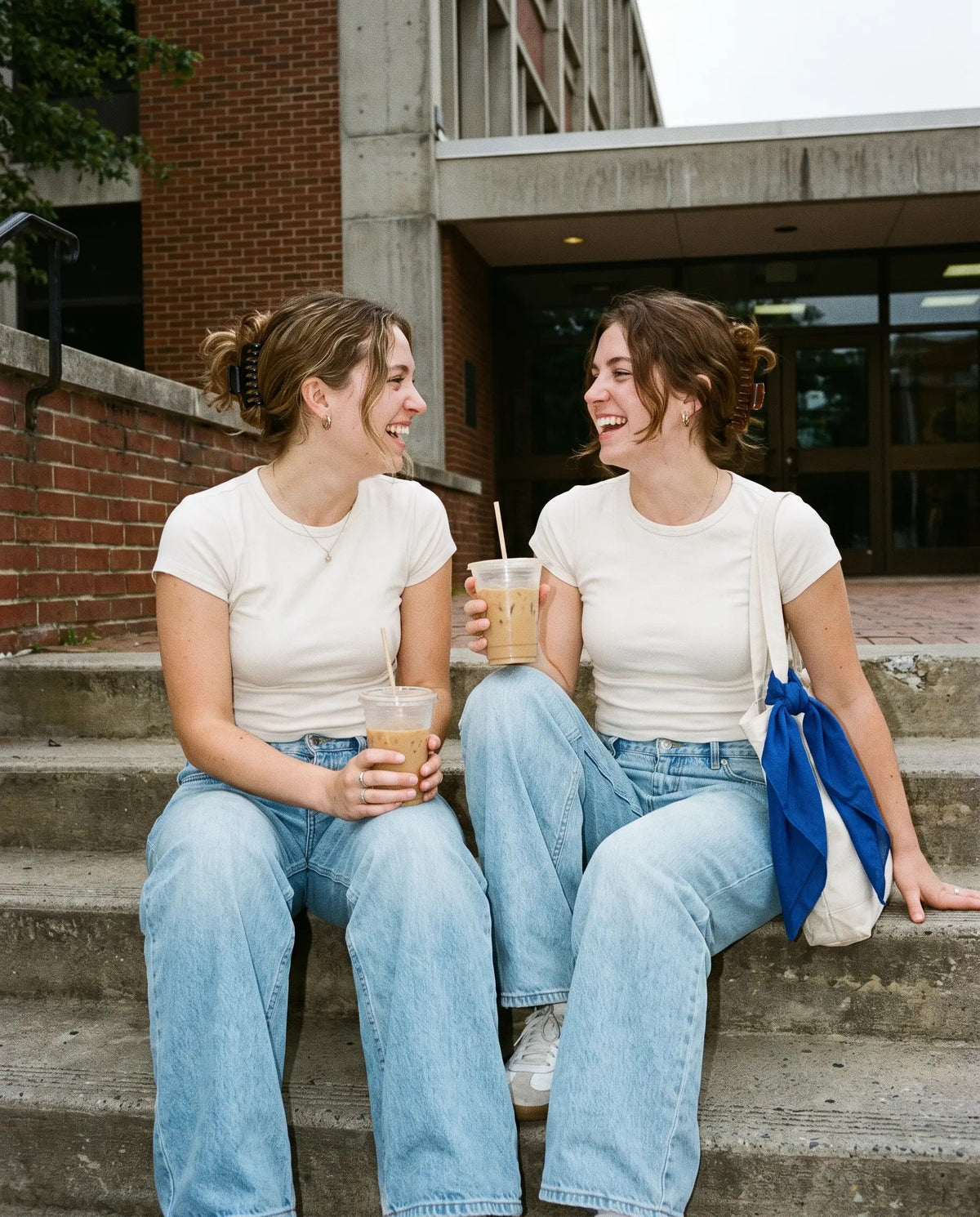 Two friends on concrete steps in white baby tees and light wide-leg jeans holding iced coffees