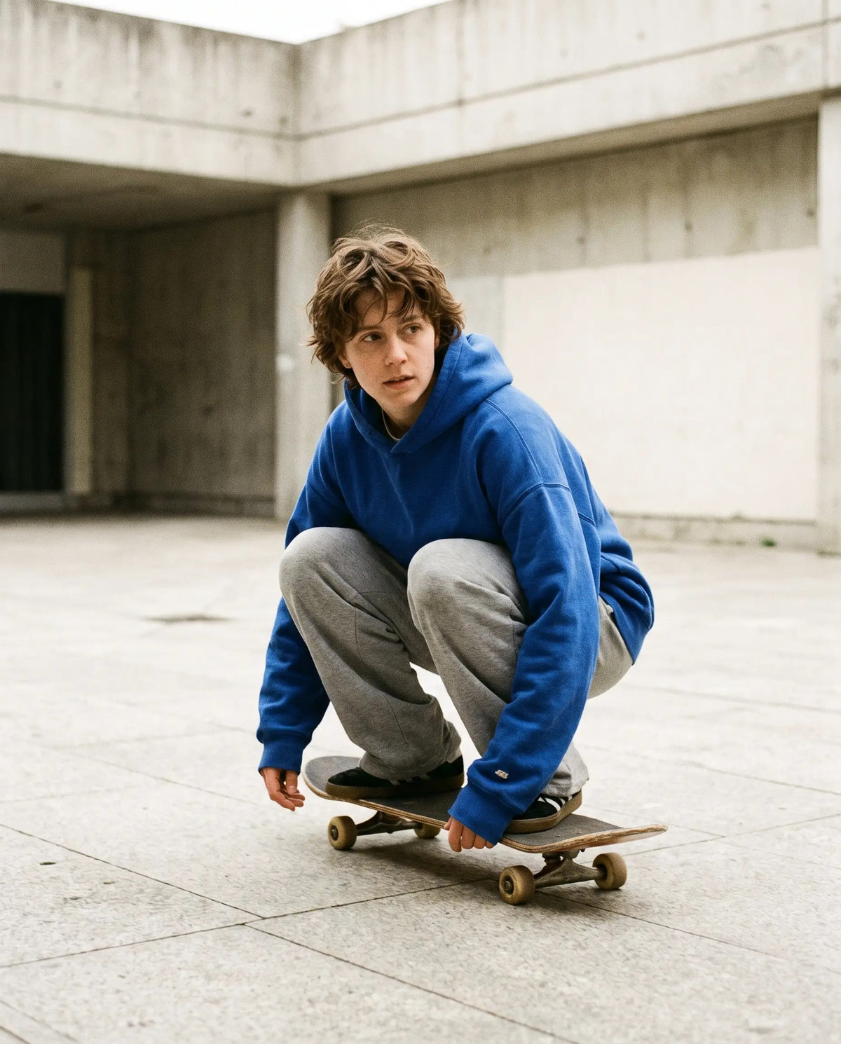 Skater crouching on a board in a cobalt hoodie and grey sweatpants in a concrete plaza
